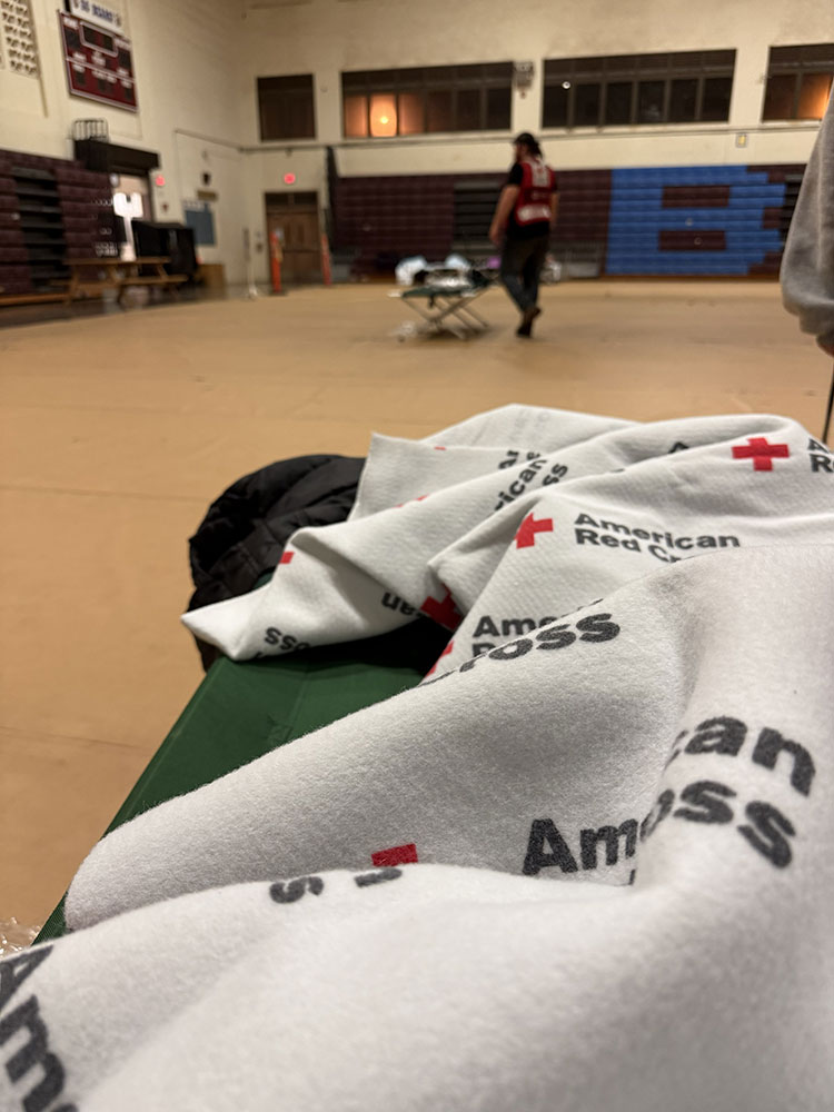 A gym converted into a shelter with an American Red Cross blanket on a cot in focus. A person in a Red Cross vest walks in the background.