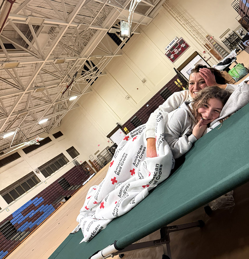 A woman and child smiling, lie on a cot covered with a Red Cross blanket in a Red Cross shelter.