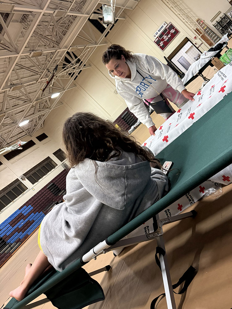 In a Red Cross shelter, a woman in a gray hoodie leans over talking to a person reclined on a cot.