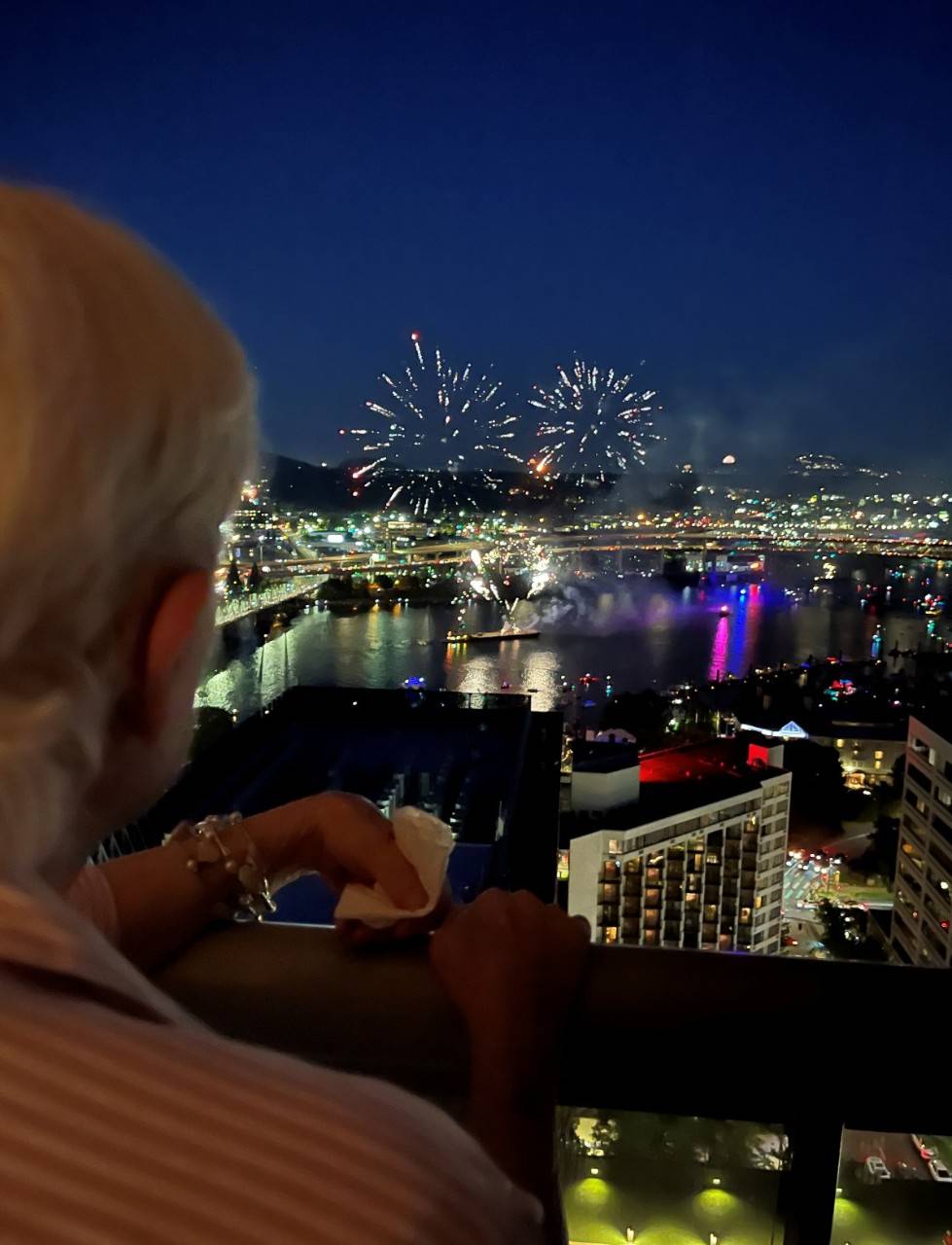 a person watching Fireworks over a harbor with boats in the water at night