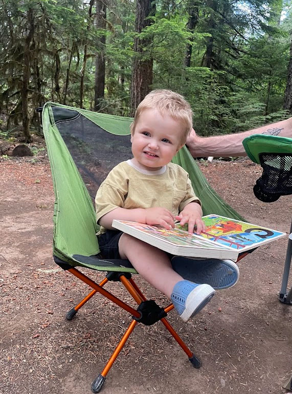 a young child sitting in a folding chair in the forest and holding a childrens book.