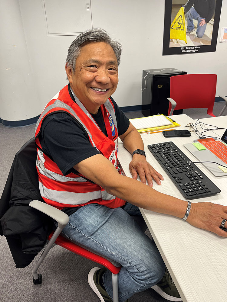 Dan Lam wearing a Red Cross vest, sitting at a desk working on a computer and smiling.
