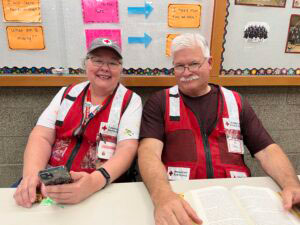 Two smiling people in Red Cross vests sit at a table, one holding a smartphone. A bulletin board with papers is visible in the background.