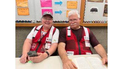 Two smiling people in Red Cross vests sit at a table, one holding a smartphone. A bulletin board with papers is visible in the background.