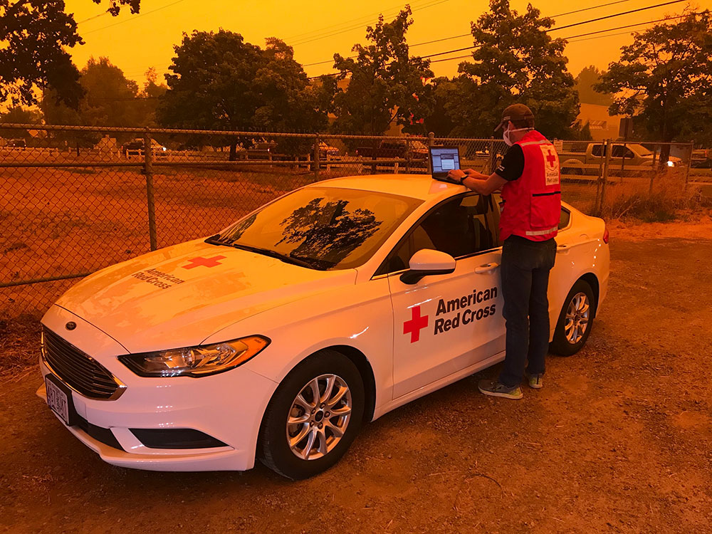 red cross volunteer wearing mask and working on a laptop on top of a red cross car.