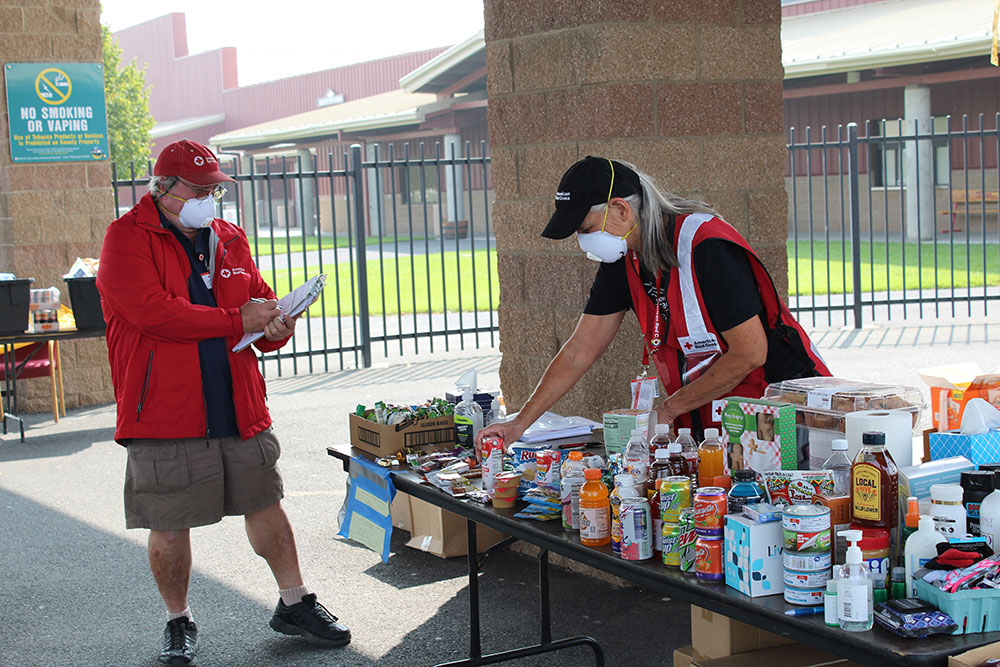 two red cross volunteers wearing masks and standing next to a table full of food, drinks and supplies.