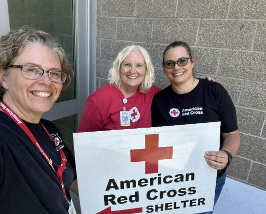 three Red Cross volunteers hold up a large American Red Cross shelter sign at the front of a building.
