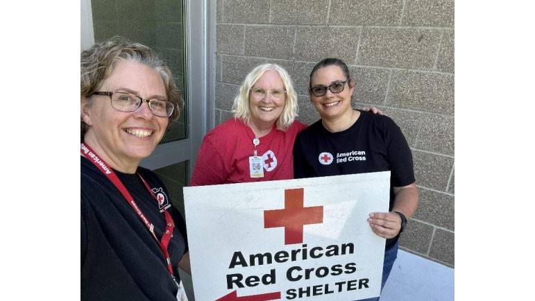 three Red Cross volunteers hold up a large American Red Cross shelter sign at the front of a building.