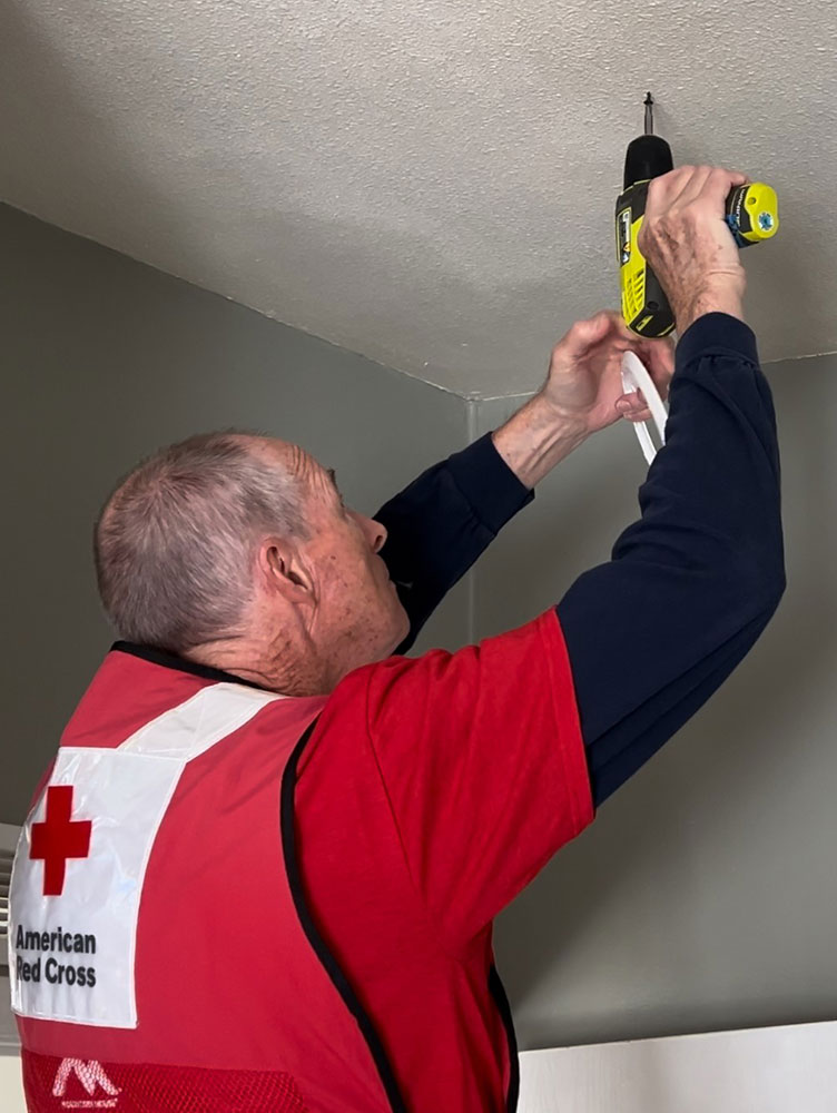 red cross volunteer holding a power drill, installing a smoke alarm on a ceiling.