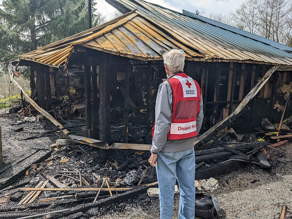 Red Cross volunteer standing next to house that has been burned.