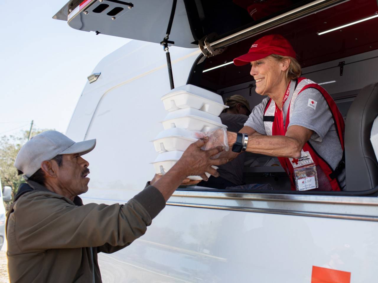 October 14, 2024. Wimauma, Florida.
American Red Cross volunteer Susan Williams serves warm meals to Cruz Salines, a resident of Wimauma, Florida. The warm meals are provided by Operation BBQ Relief, a community partner, and are served alongside water and snacks. The Wimauma community has been without power, and the food served by volunteers was frequently the first warm meal many residents enjoyed in several days. The Red Cross has hundreds of volunteers on the ground in communities working alongside community partners and government agencies to provide much-needed services to those who were impacted by both Hurricane Helene and Hurricane Milton. 
Photo by Marko Kokic/American Red Cross

