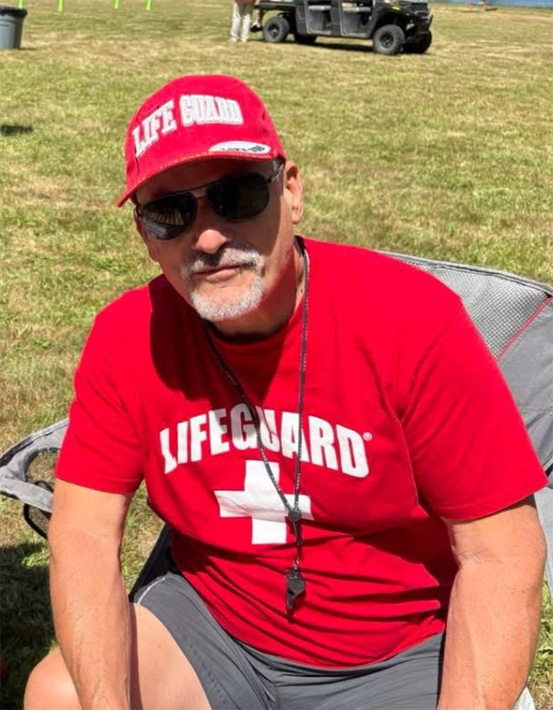 Greg Kaminski wearing sunglasses and a life guard hat and shirt while sitting in a lawn chair.