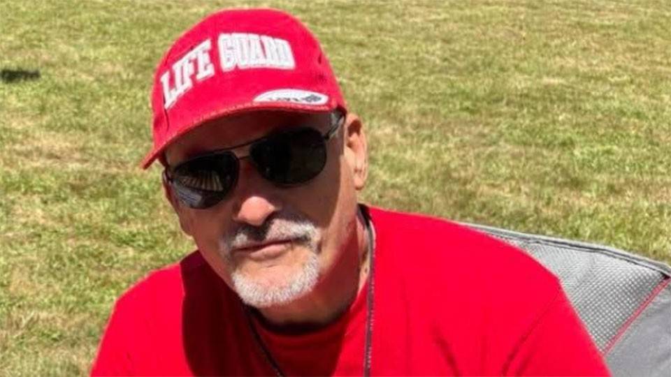 Greg Kaminski wearing sunglasses and a life guard hat and shirt while sitting in a lawn chair.