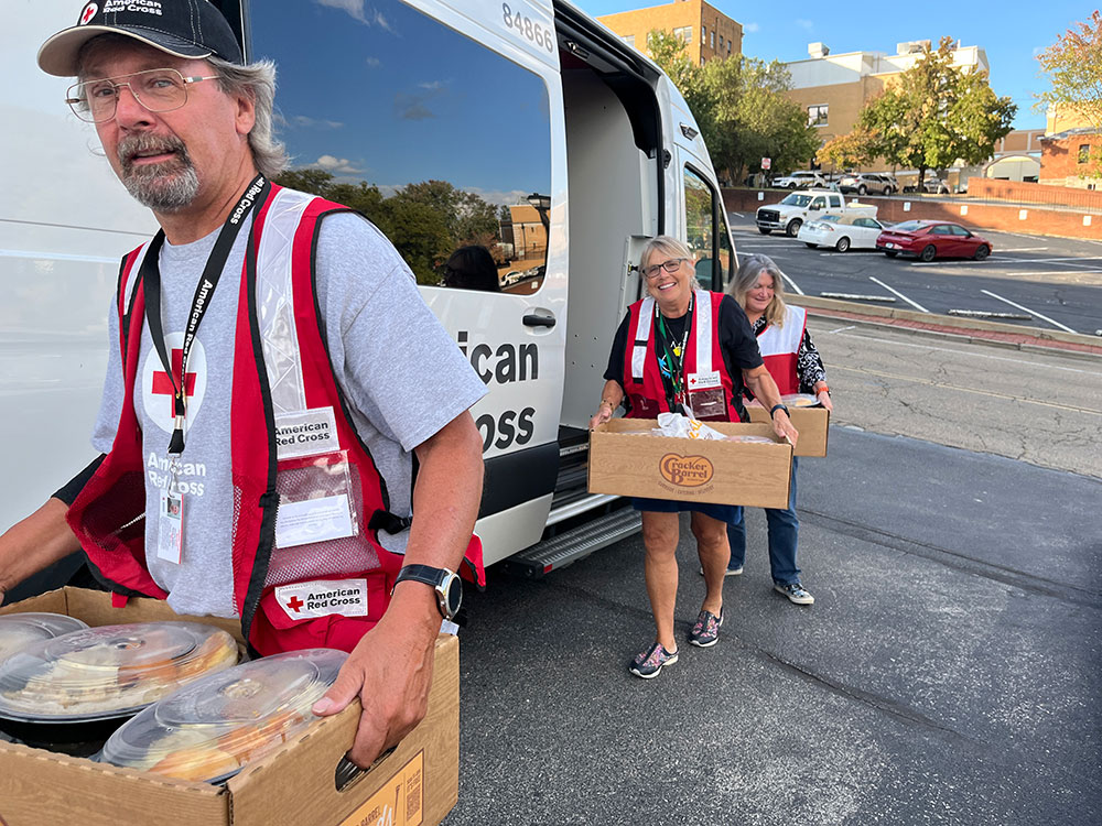 red cross volunteers carrying food in boxes from a red cross van.