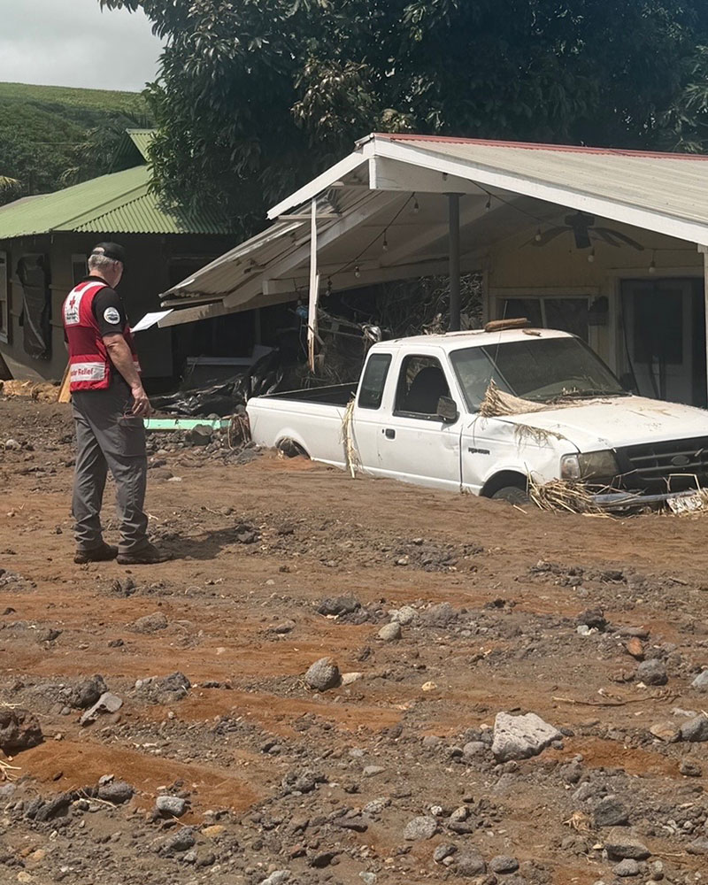 a Red Cross volunteer standing next to a house and a truck that's buried halfway in dirt