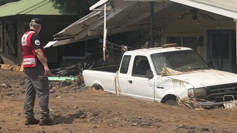 a Red Cross volunteer standing next to a house and a truck that's buried halfway in dirt