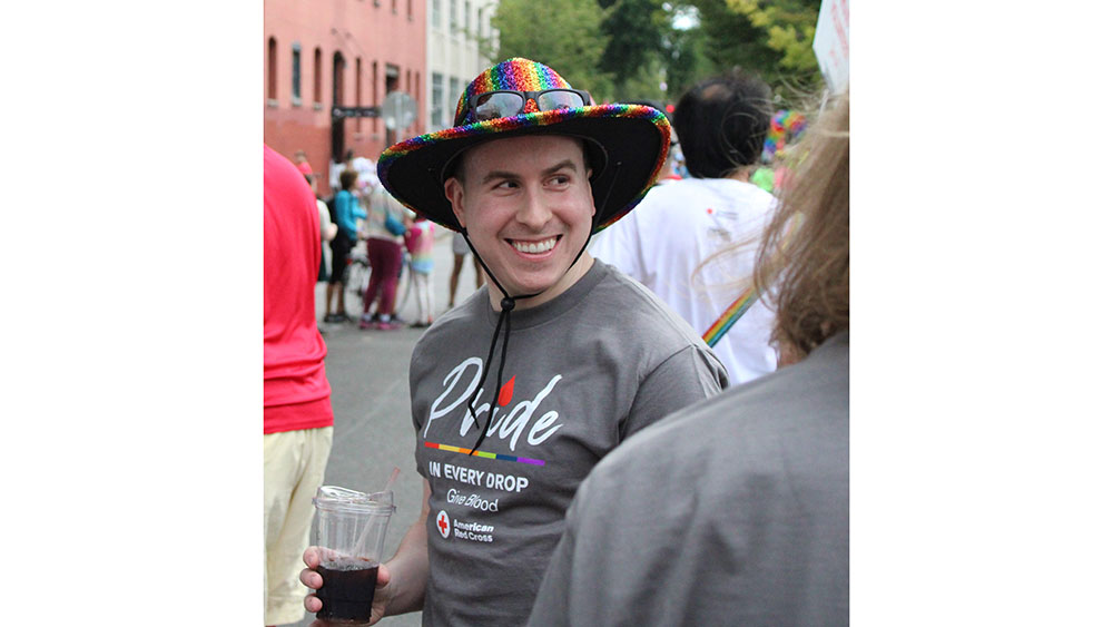 Person at the Pride parade holding a drink and wearing a rainbow hat and red cross pride shirt.
