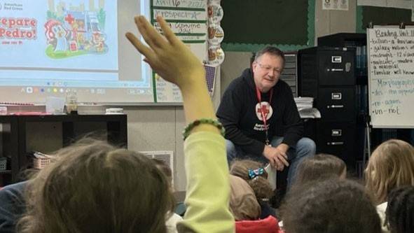 A classroom setting with Jens Haetty sitting on a table, engaging a group of children. One child in the foreground raises a hand. A projector displays a colorful slide titled "Prepare with Pedro."