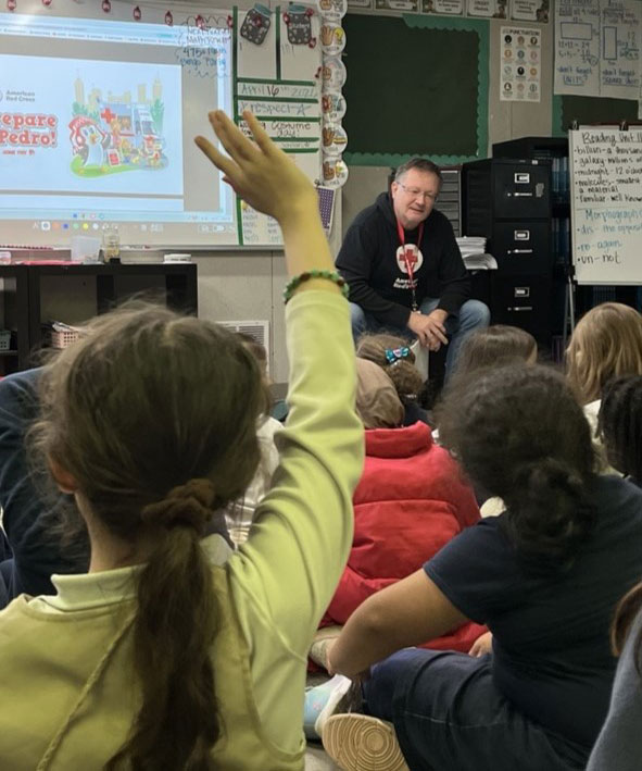 A classroom setting with Jens Haetty sitting on a table, engaging a group of children. One child in the foreground raises a hand. A projector displays a colorful slide titled "Prepare with Pedro."