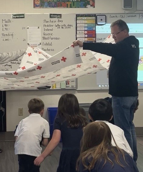 Jens Haetty holds a Red Cross emergency blanket as children crouch beneath it in a classroom. Whiteboard with lessons and colorful decor visible.