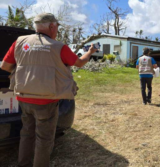  Jim Court and another Red Crosser in Red Cross vests stands in front of a damaged house.