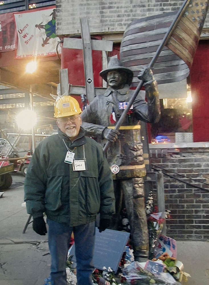 Jim Stevenson, wearing a hard hat, glasses, jacket and gloves, standing next to a firefighter statue that's holding an American flag.
