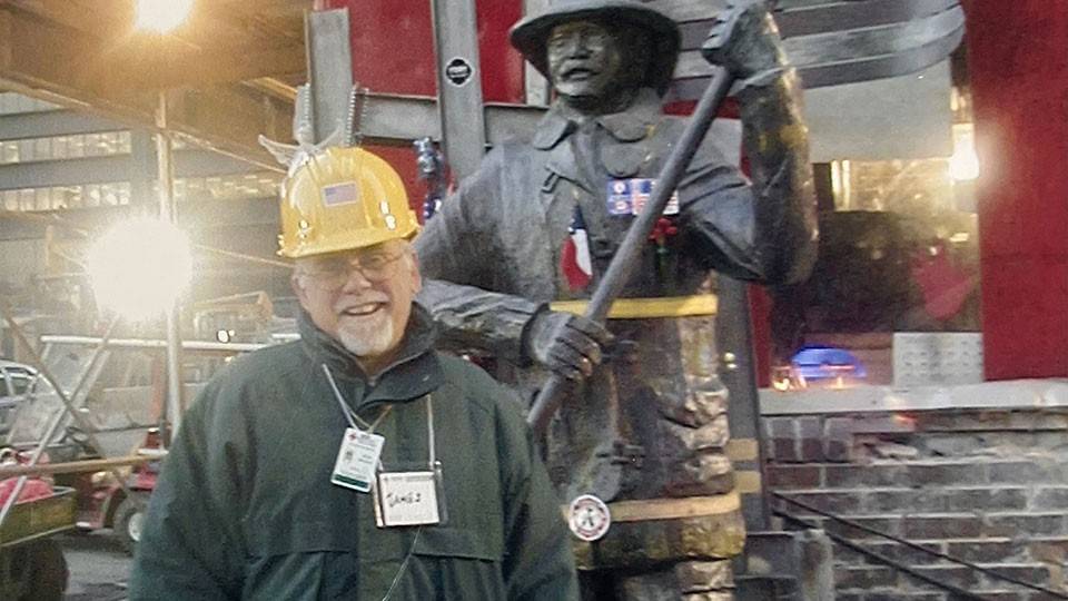 Jim Stevenson, wearing a hard hat, glasses, jacket and gloves, standing next to a firefighter statue that's holding an American flag.