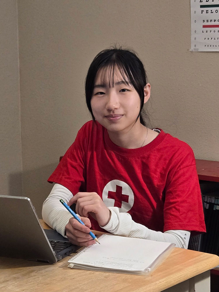 Katherine Liu, wearing a Red Cross shirt, sitting at a desk with a laptop and notebook.