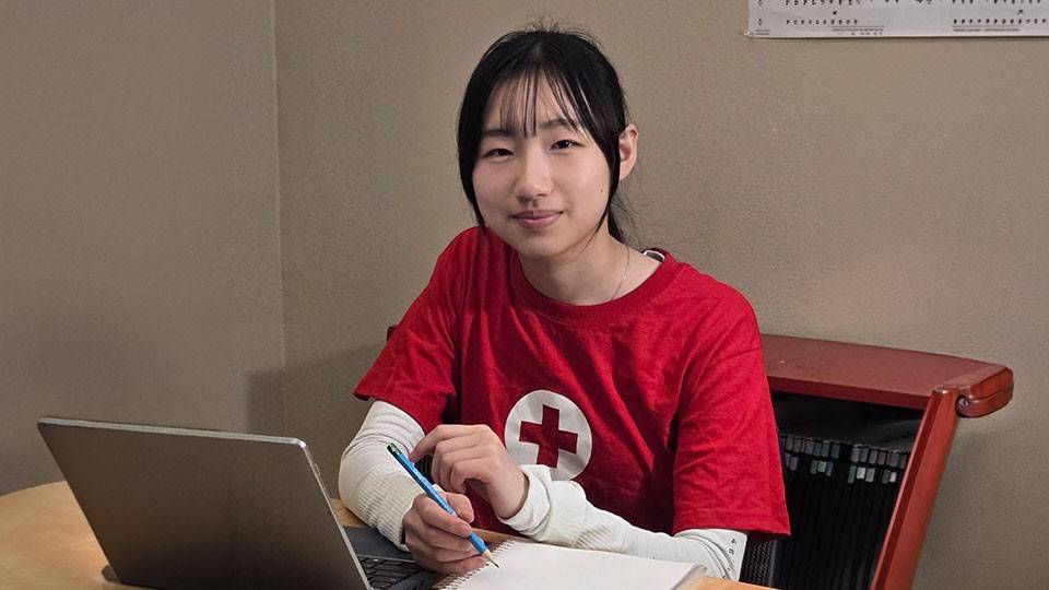 Katherine Liu, wearing a Red Cross shirt, sitting at a desk with a laptop and notebook.
