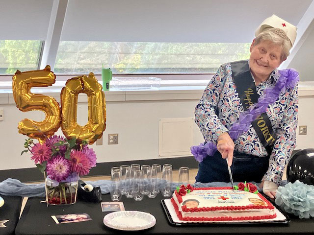 Marcia Raisanen next to a 50 balloon wearing a Red Cross nurse hat and a sash while cutting a cake. 