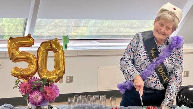 Marcia Raisanen next to a 50 balloon wearing a Red Cross nurse hat and a sash while cutting a cake. 