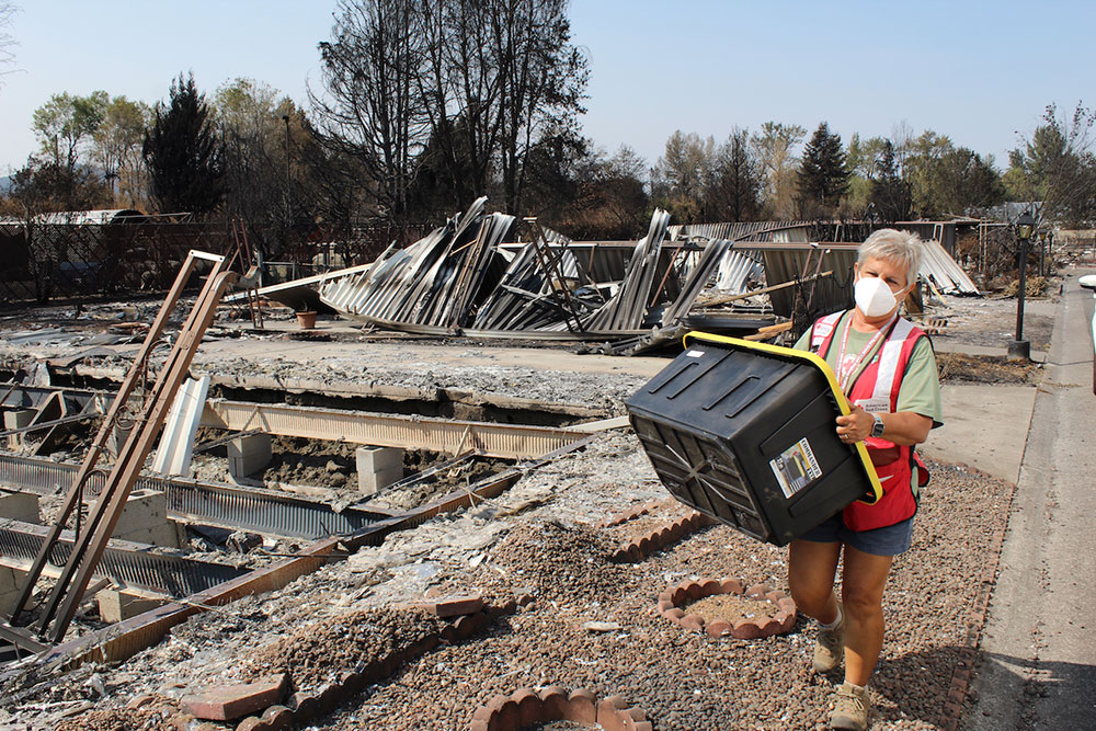 red cross volunteer wearing mask and carrying a storage container while walking past a burned down house.