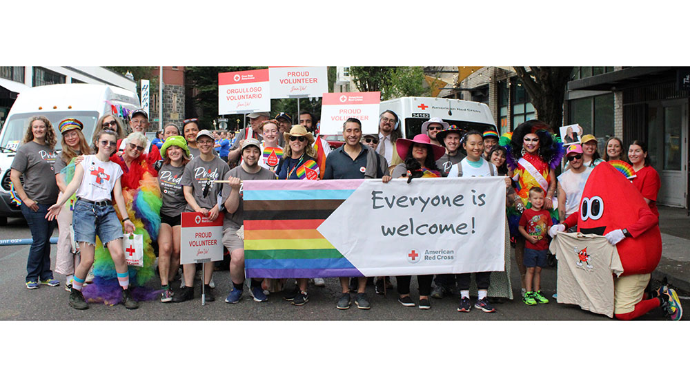 group of people and red cross blood drop mascot pose for pic in Pride parade while holding a large Everyone is welcome! banner.