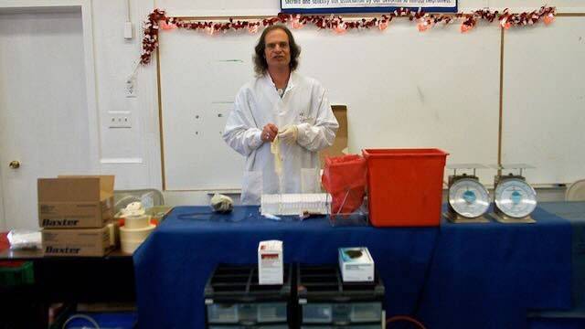 Robert Cork, wearing a lab coat and rubber gloves, standing behind a table with scales and other items.