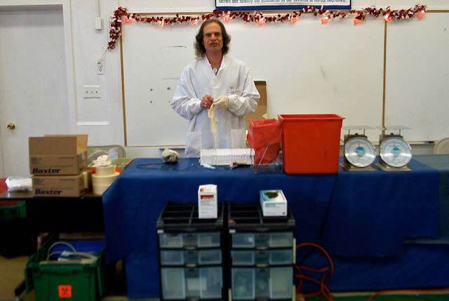 Robert Cork, wearing a lab coat and rubber gloves, standing behind a table with scales and other items.