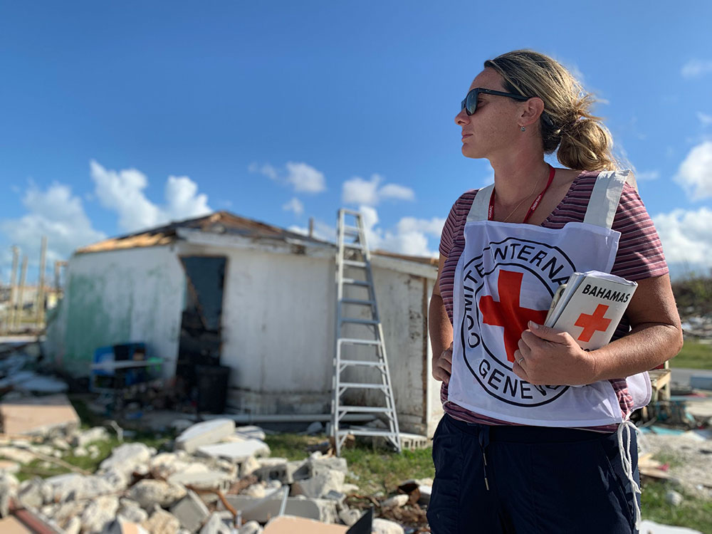 A woman in a red cross shirt stands before a destroyed house.