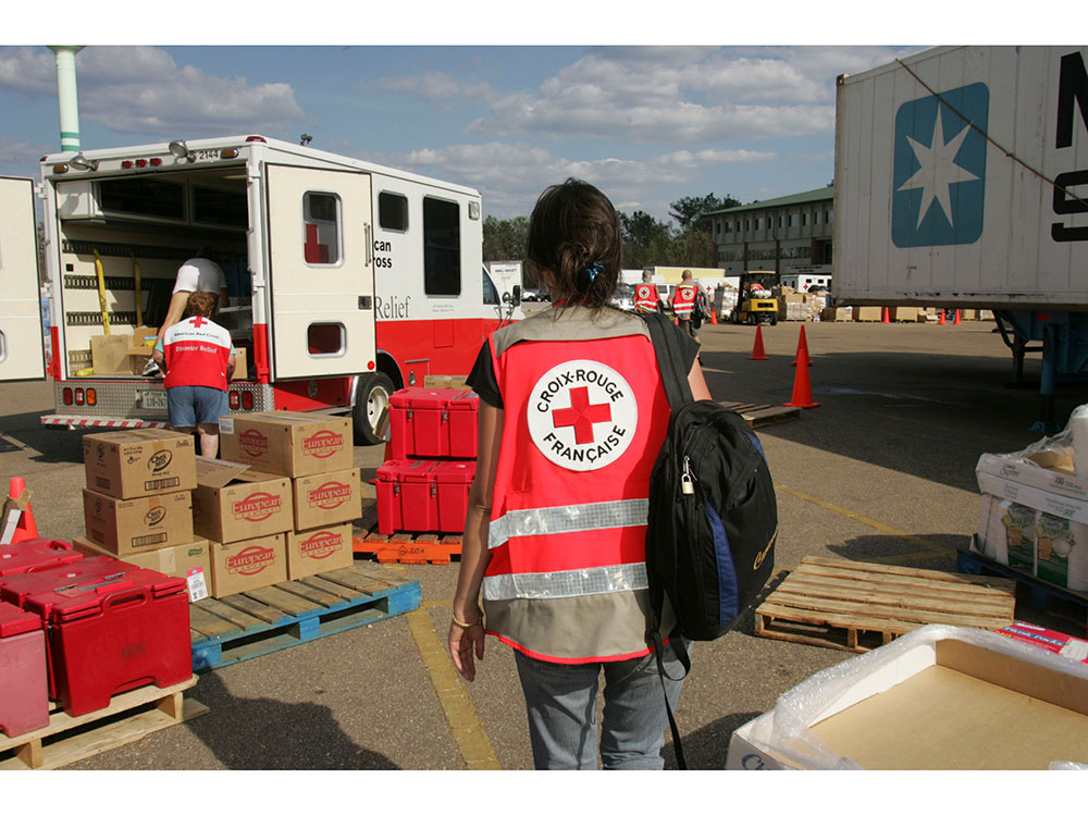 A woman in a Red Cross vest walks towards a parked Red Cross truck to help with unloading boxes.