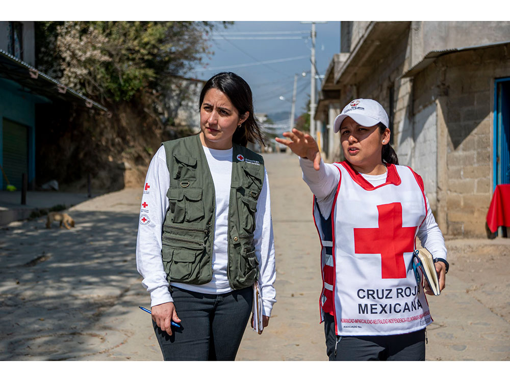 Two women dressed in Red Cross shirts and white vests stand on a street, engaged in conversation.