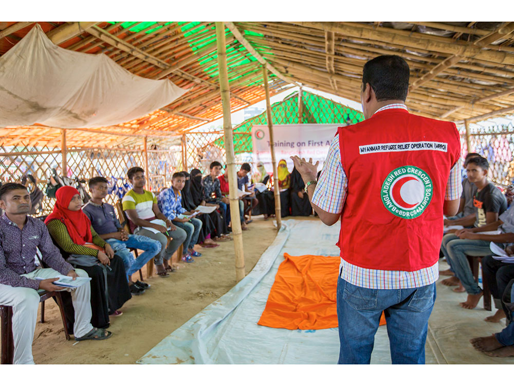 A man wearing a red vest delivers a presentation to a captivated group of listeners in a conference setting.