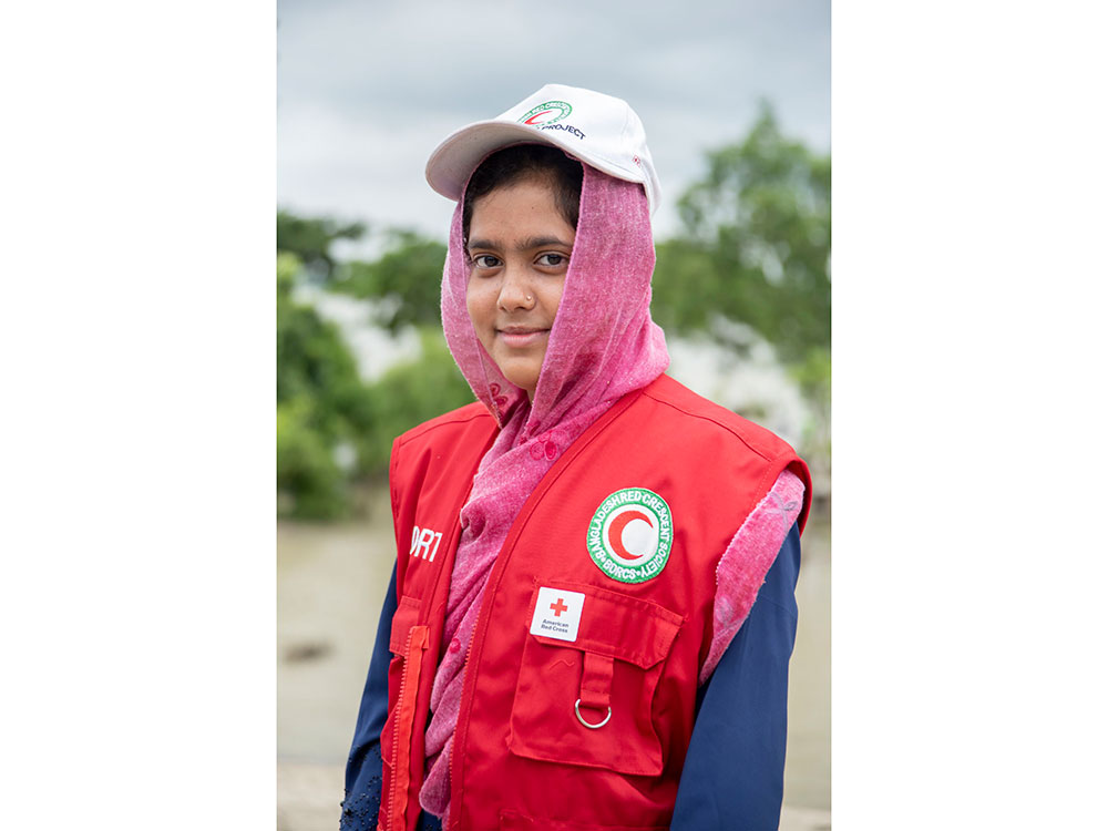 A young woman in a red vest and white hat smiles, standing outdoors against a blurred background.