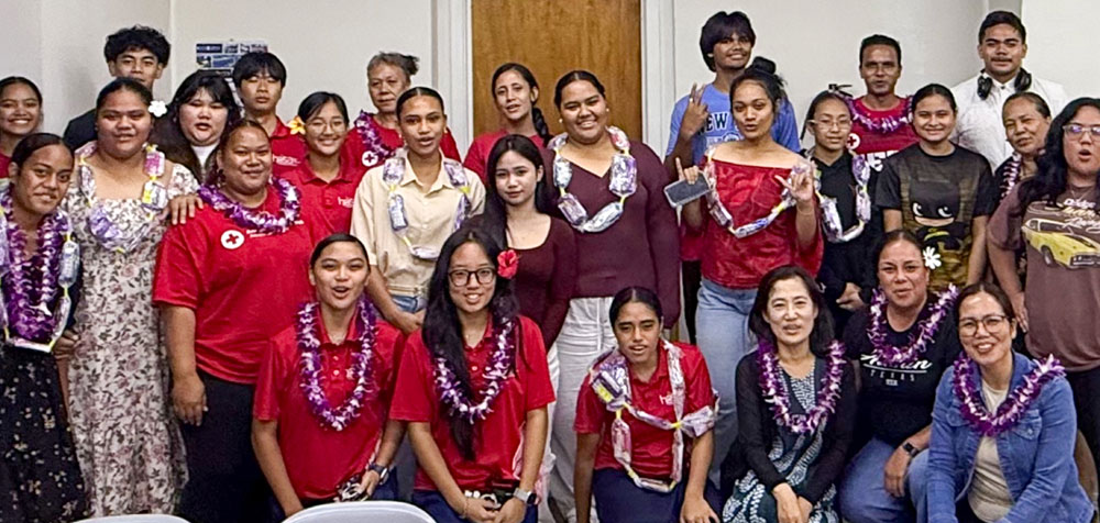 A group of people smiling and posing in a room. Many wear purple leis and red cross shirts.