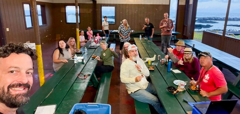 Group of people gathered around green picnic tables inside a wooden pavilion. They are smiling and appear to be enjoying food together, with an ocean view in the background.
