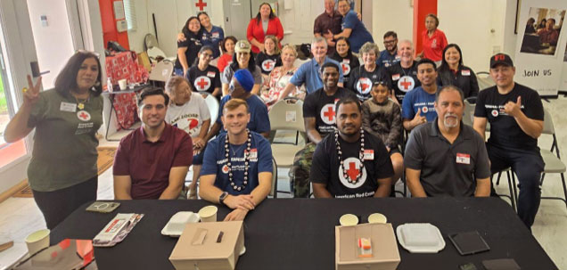 Group of red cross volunteers, many wearing Red Cross shirts, smiling, and seated at tables with food containers.