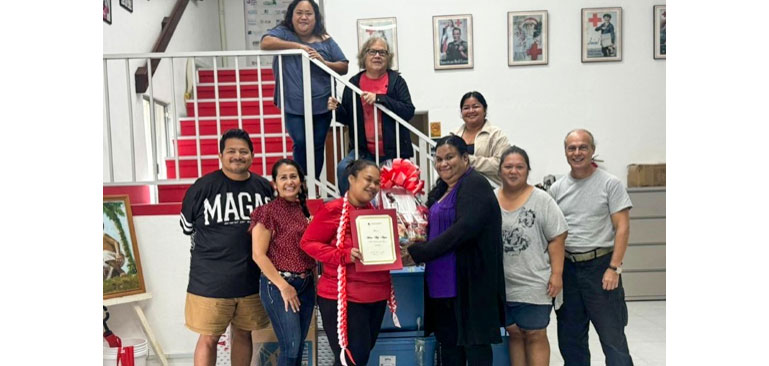 A group of nine people poses indoors, smiling and holding a framed red cross certificate and a gift basket with red ribbons.
