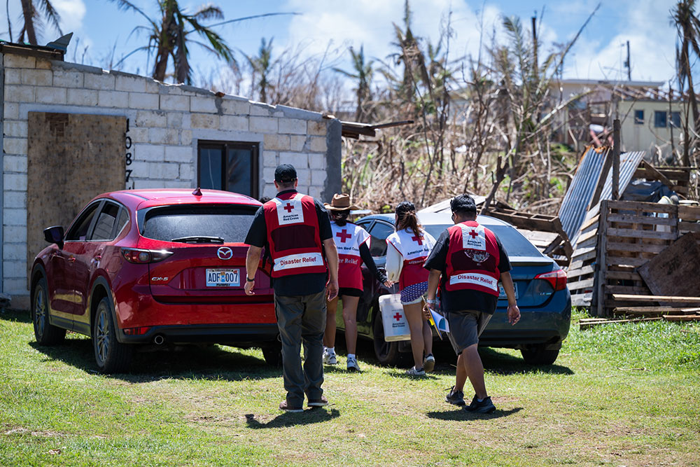Red Cross workers in disaster relief vests walk toward a damaged building holding supplies. A red car and debris are visible.