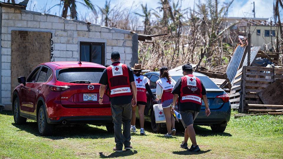 Red Cross workers in disaster relief vests walk toward a damaged building holding supplies. A red car and debris are visible.
