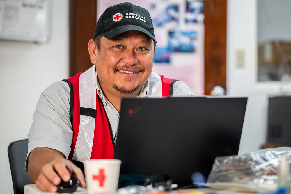 JD Tenorio in a Red Cross cap and vest works on a laptop. Coffee cup with Red Cross logo in foreground.