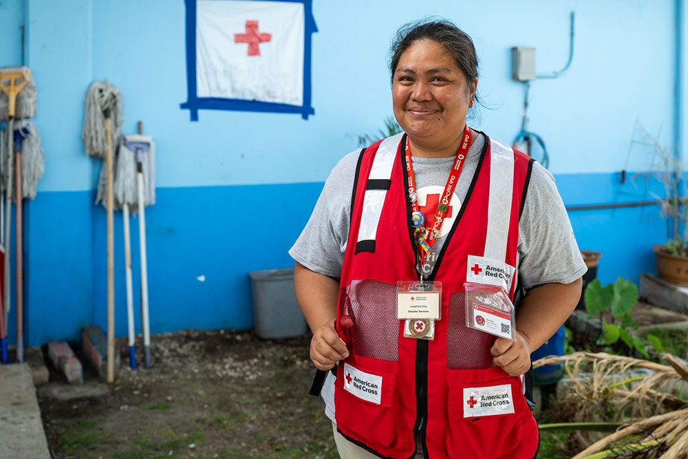 Zoe King smiling in a Red Cross vest stands in a room with blue walls, mops and plants in the background.