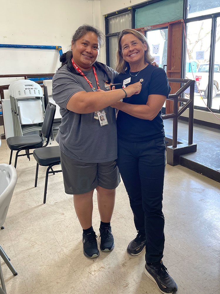 Two people indoors, each pointing to a Red Cross button on the other’s shirt; stacked chairs and a window in the background on a linoleum floor.