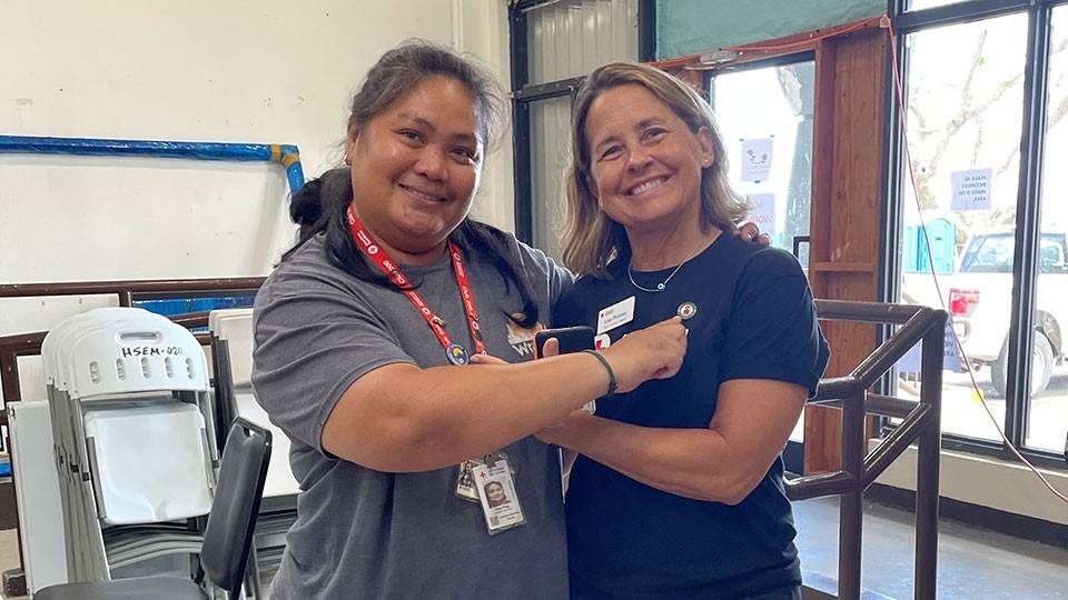 Two people indoors, each pointing to a Red Cross button on the other’s shirt; stacked chairs and a window in the background on a linoleum floor.